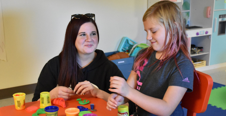 An adult and child smile while playing with Play-Doh in a colorful classroom environment.